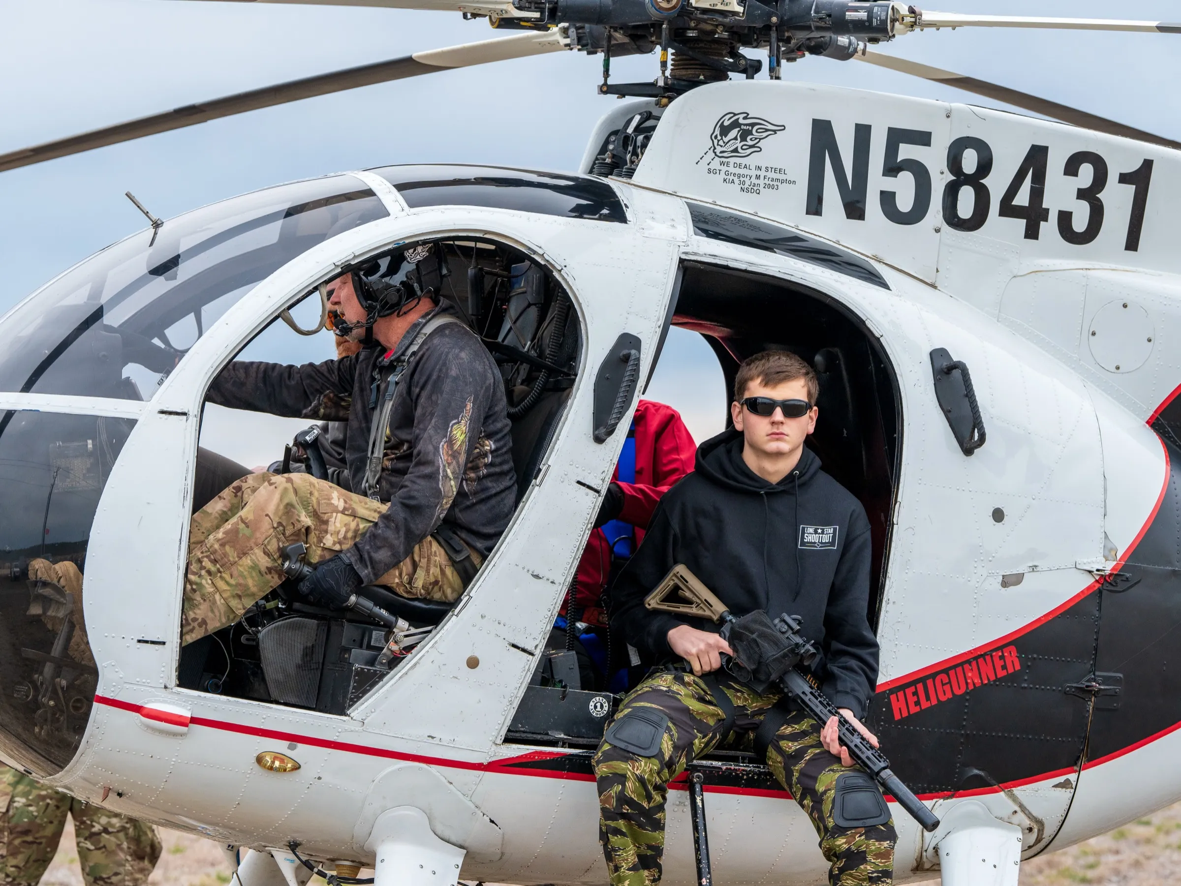Shooter in a Lone Star Shootout hoodie seated in the Heligunner helicopter door with a rifle, pilot at the controls